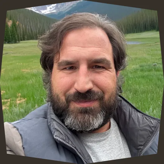 Aaron smiling outdoors with mountains and a meadow behind him